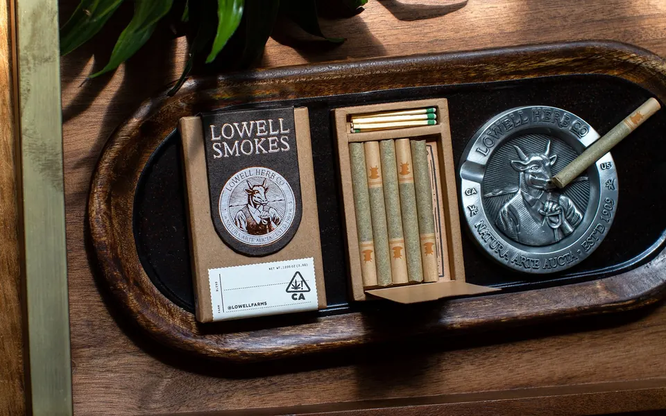 A Lowell Smokes pack, its open tray of pre-rolls and the Signature Metal Ashtray on a wooden valet tray